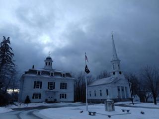 A white two story building next to a white church with steeple with snow on the ground and a cloudy sky.